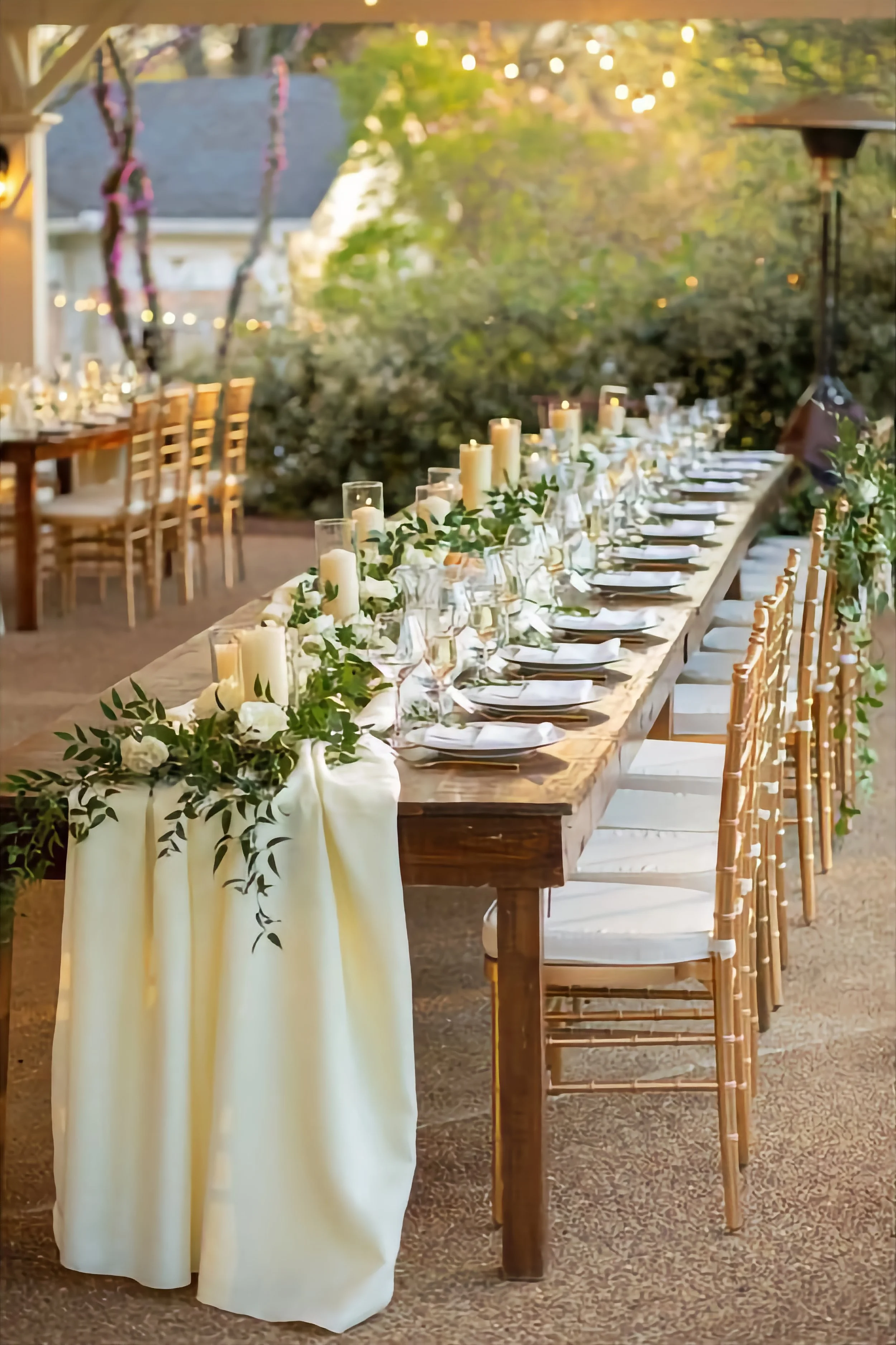 Elegant outdoor banquet table decorated with candles, green foliage, white flowers, white tablecloths, and gold chairs, set up for evening dining in a garden with string lights above.
