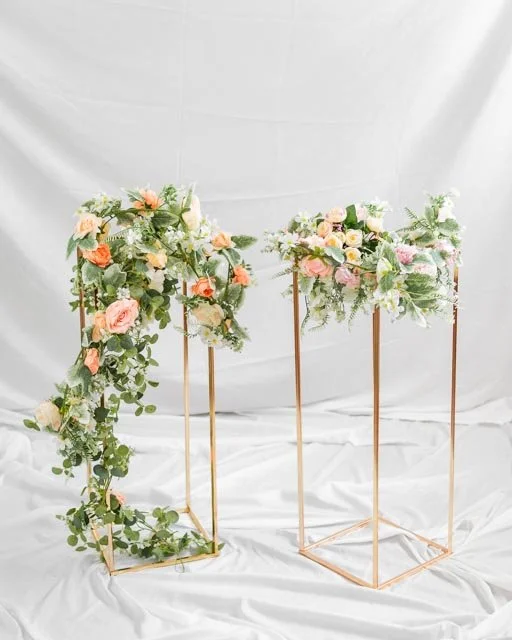 Two gold metal stands with floral arrangements featuring pink and white roses and green foliage against a white background.
