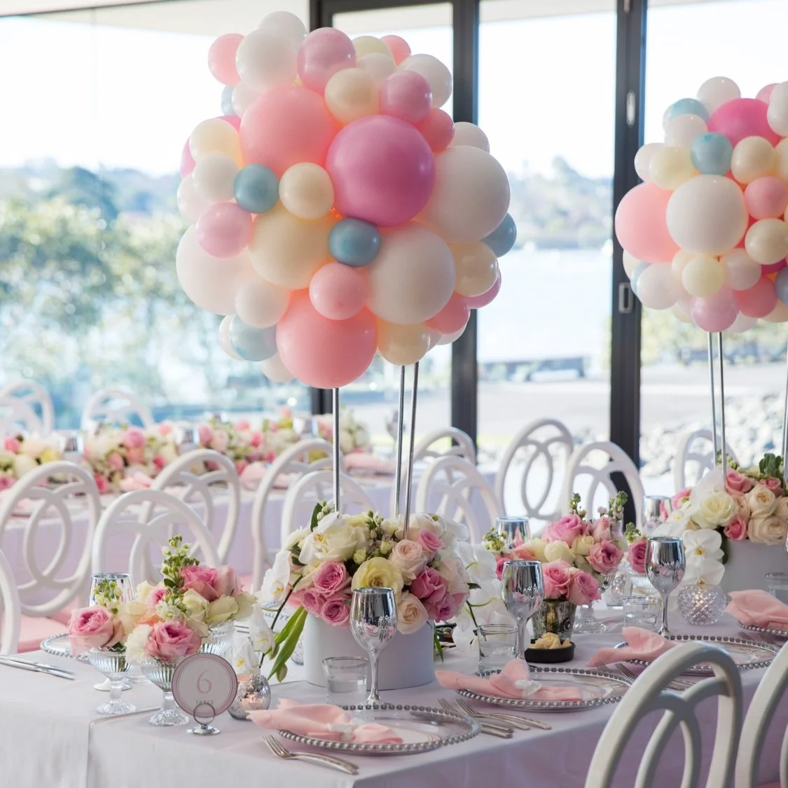 Elegant event table setting with white, pink, blue, and cream balloon centerpieces, floral arrangements, and tableware.