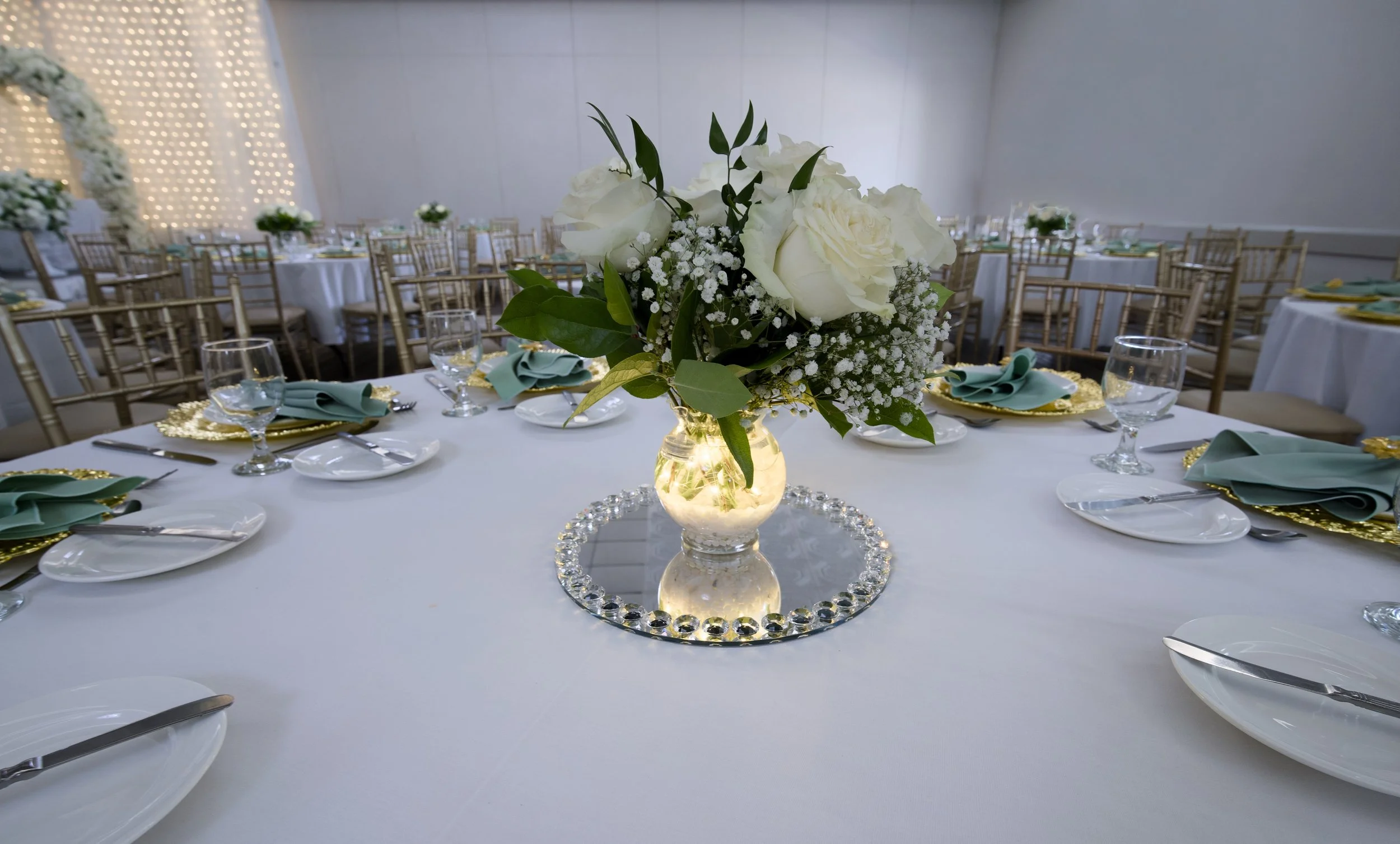 Wedding reception table setting with white roses and baby's breath centerpiece on mirrored base, surrounded by elegant place settings with glassware, golden chargers, and folded napkins.