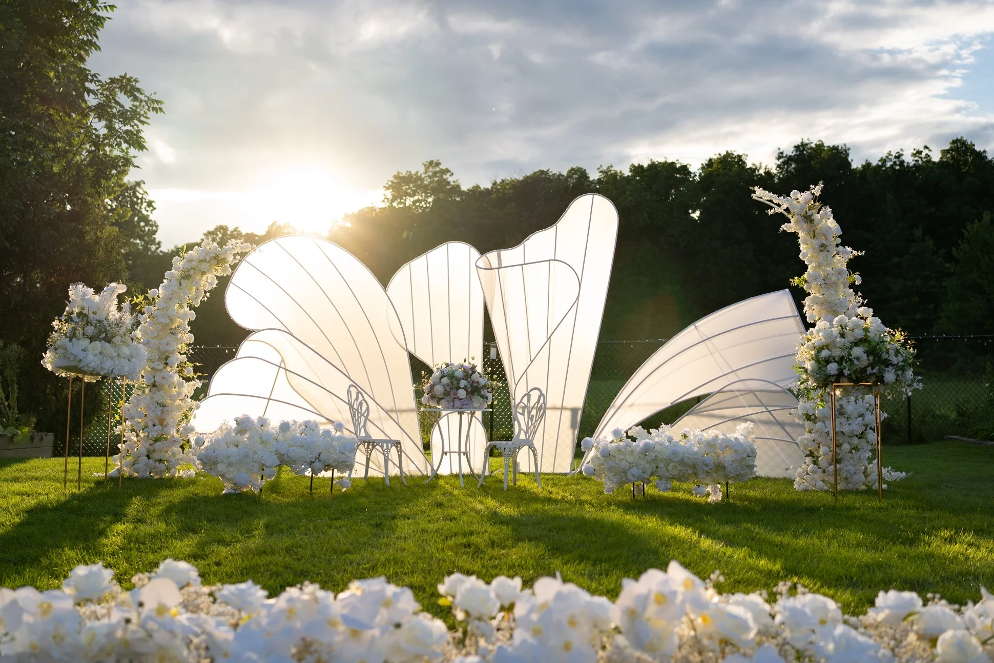 Outdoor wedding setup with white floral arrangements, abstract backdrop, and decorative chairs on a grassy field at sunset.
