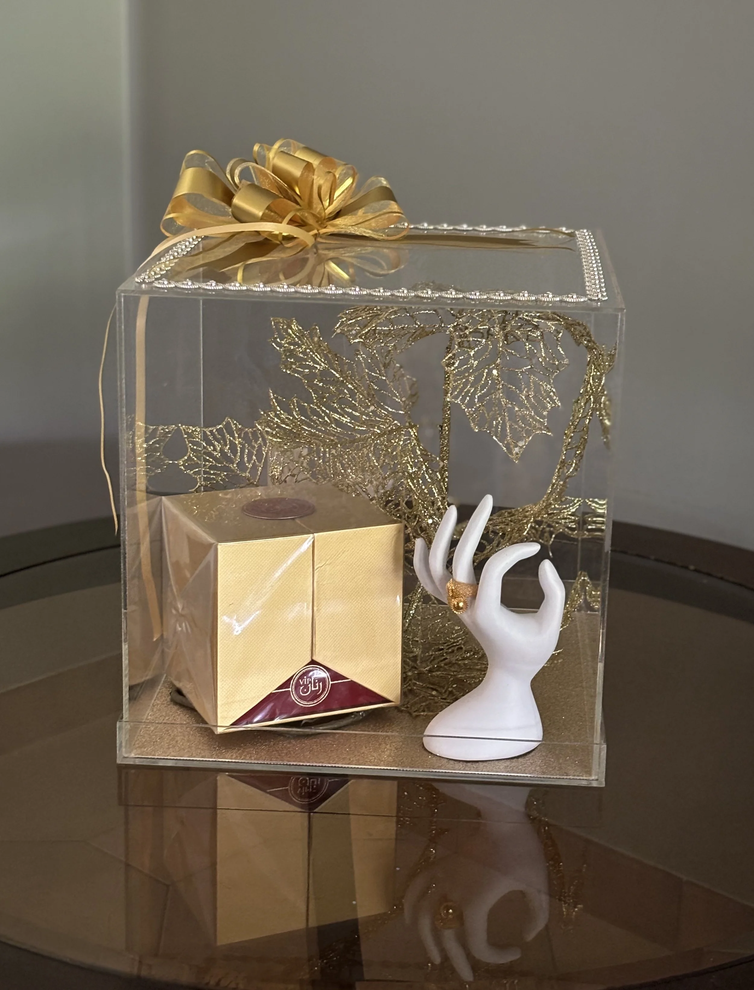 Decorative gift box with gold bow containing a gold-wrapped package and a white hand sculpture displaying a gold ring, placed on a glass table.