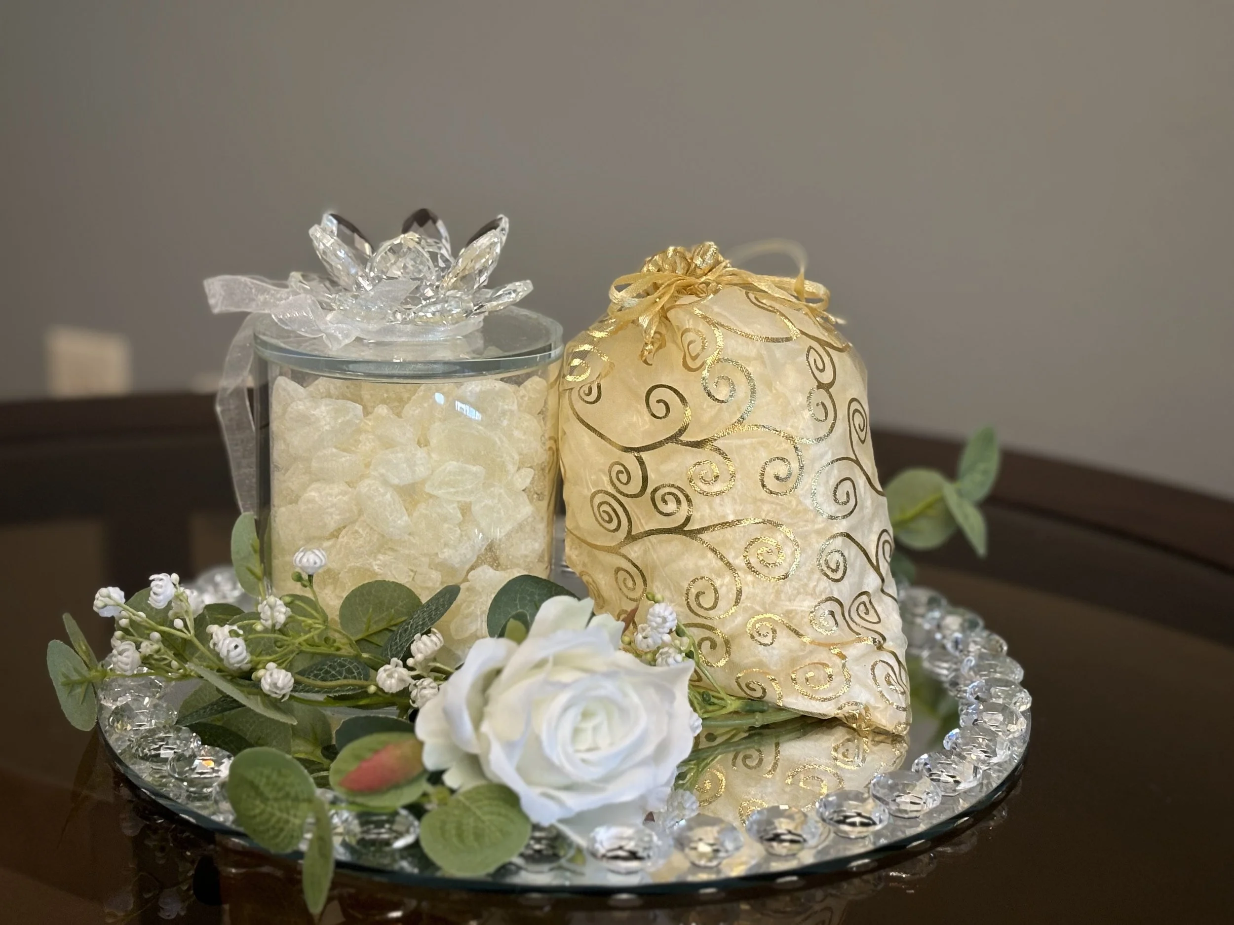 Glass jar with transparent decorative stones and a gold, patterned bag on a mirrored tray adorned with artificial flowers and leaves.
