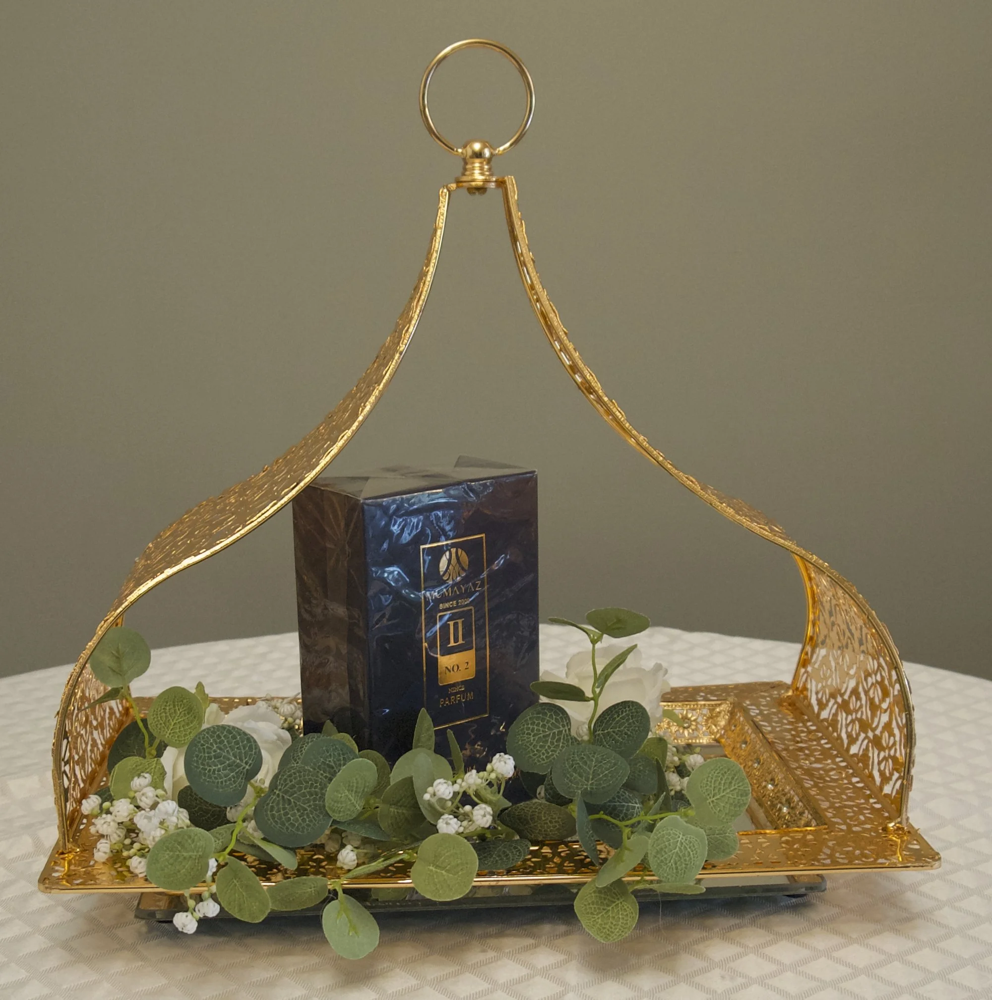Decorative gold tray with handle holding a perfume box, surrounded by artificial greenery and white flowers, displayed on a patterned tablecloth.