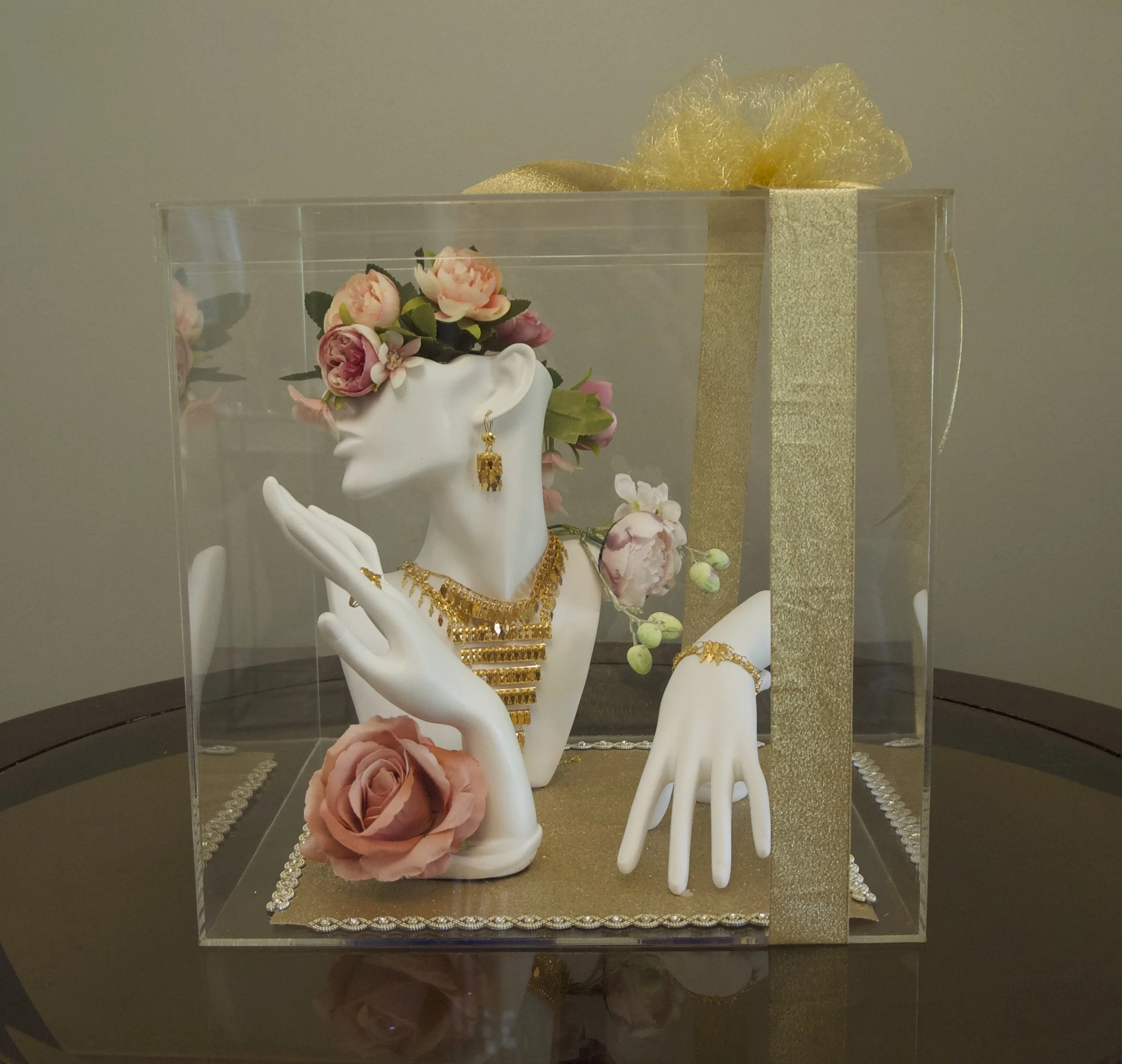 Jewelry display with a mannequin head decorated with pink roses, adorned with gold necklaces, earrings, and a bracelet, inside a clear box with gold ribbon and roses around it.