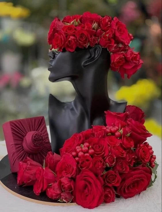 Black mannequin bust with a floral crown of red roses and a bunch of red roses in front, along with a red decorative item, all placed on a round black surface.