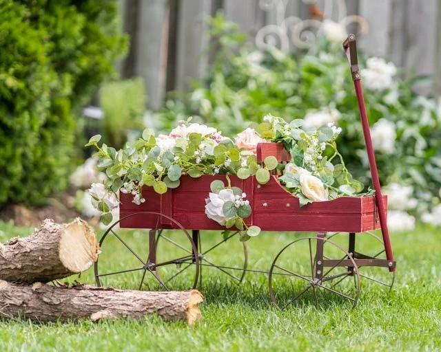 Red vintage toy wheelbarrow filled with white and pink flowers on green lawn, background of garden and trees.