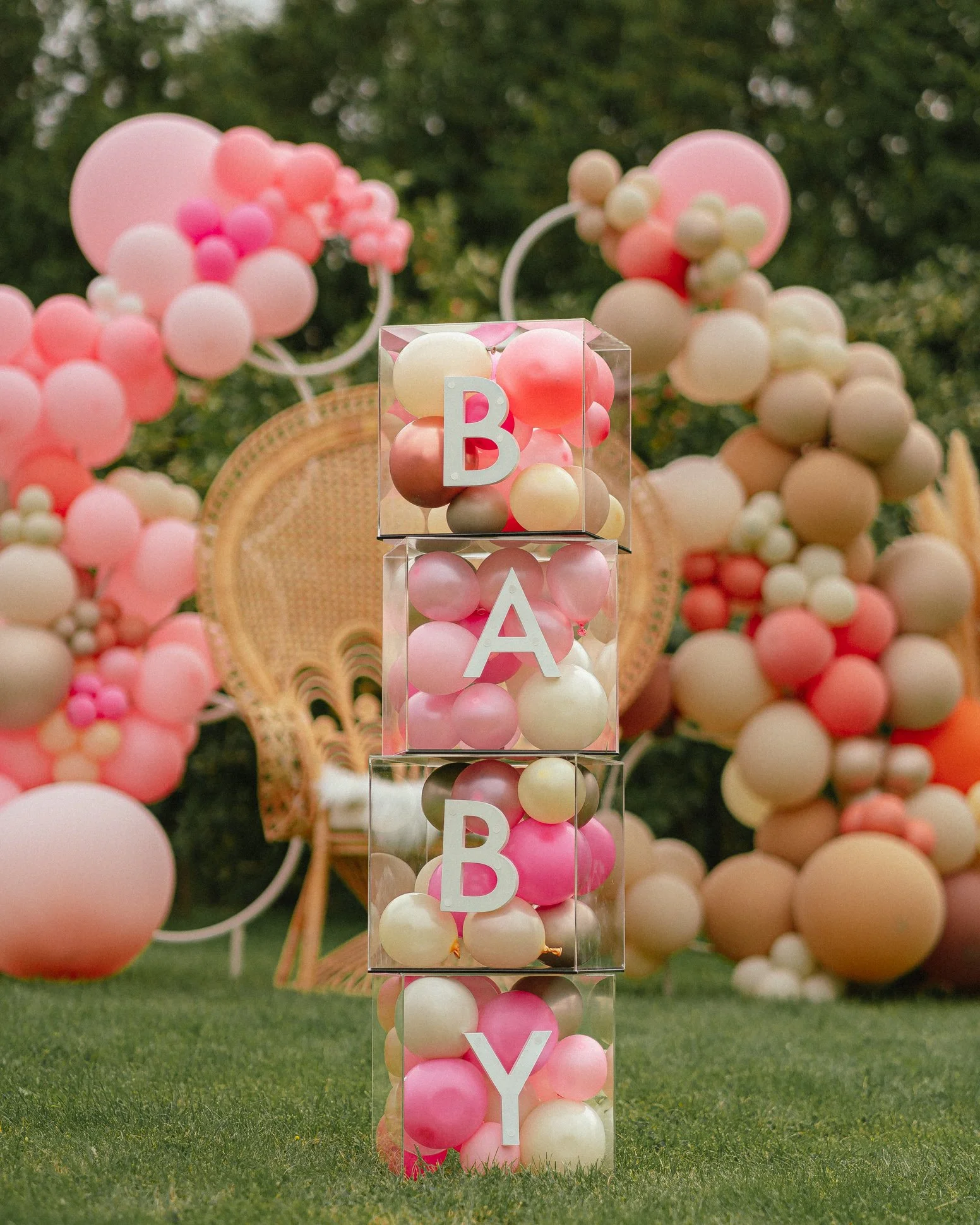 Clear blocks filled with pink, white, and peach balloons stacked to spell "BABY," against a backdrop of balloon arches and wicker chair in a garden setting.
