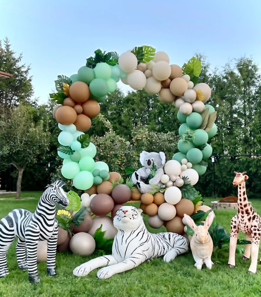 Decorative outdoor display with a balloon arch in shades of white, tan, green, and brown, featuring tropical leaves, and stuffed animal figures of a zebra, a white tiger, a rabbit, and a giraffe, set on grass with trees in the background.
