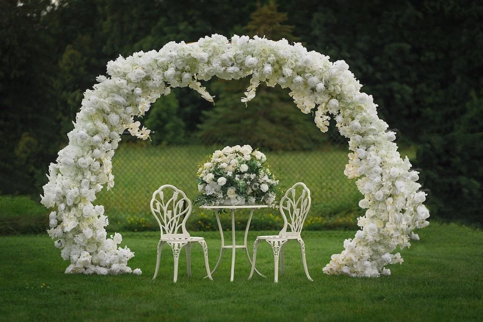 A decorative white floral arch with a small table and two chairs underneath, set outdoors on a grassy area for a wedding or event.