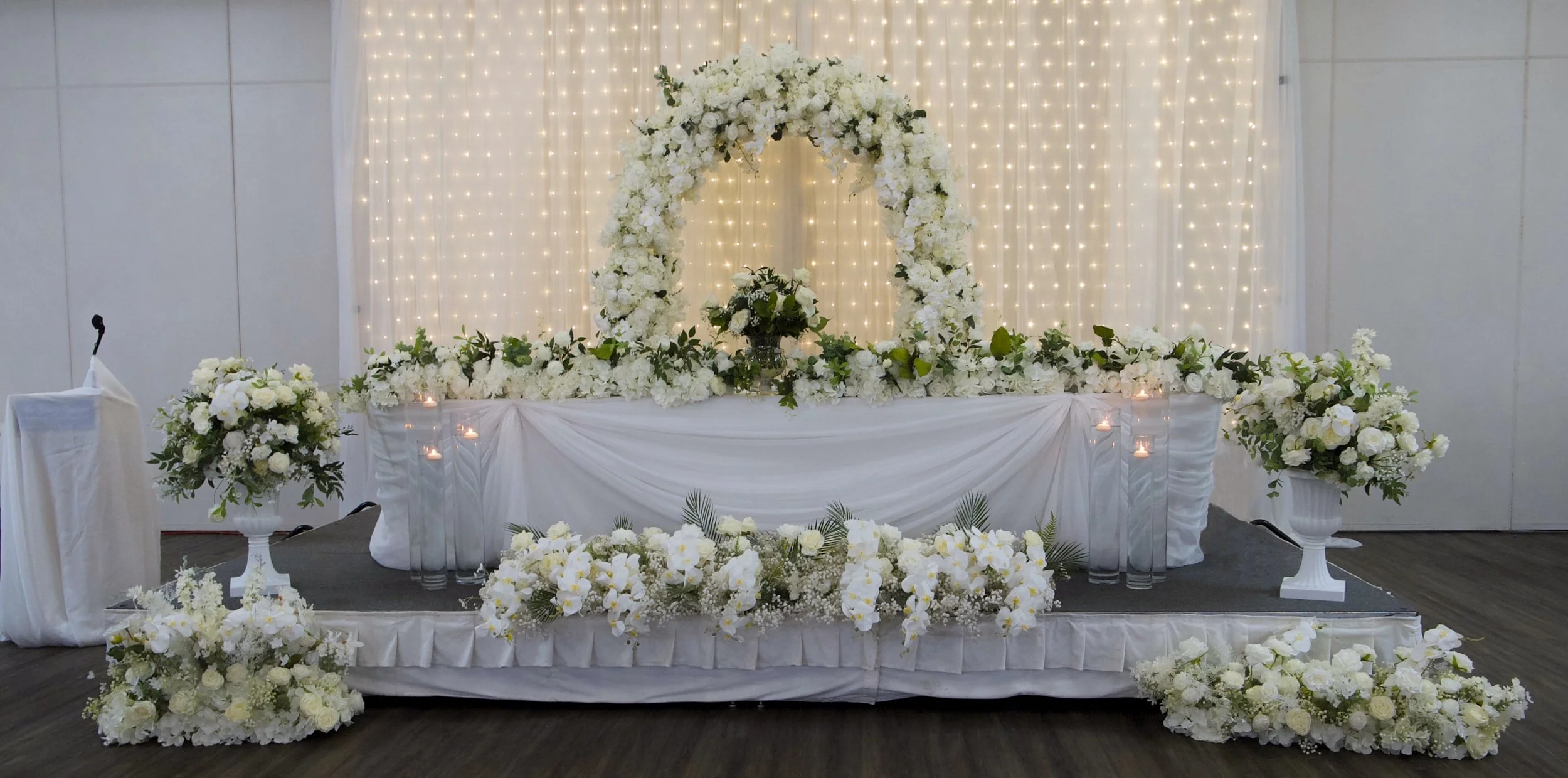 Elegant event stage decorated with white flowers, floral arch, candles, and draped fabric.