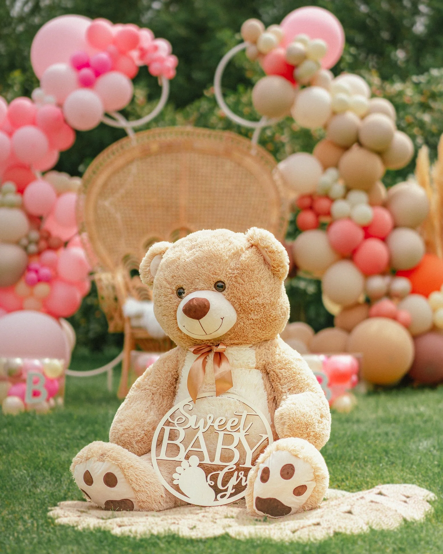 Large teddy bear with "Sweet Baby" sign, surrounded by pink and beige balloons, outdoor baby shower setup.