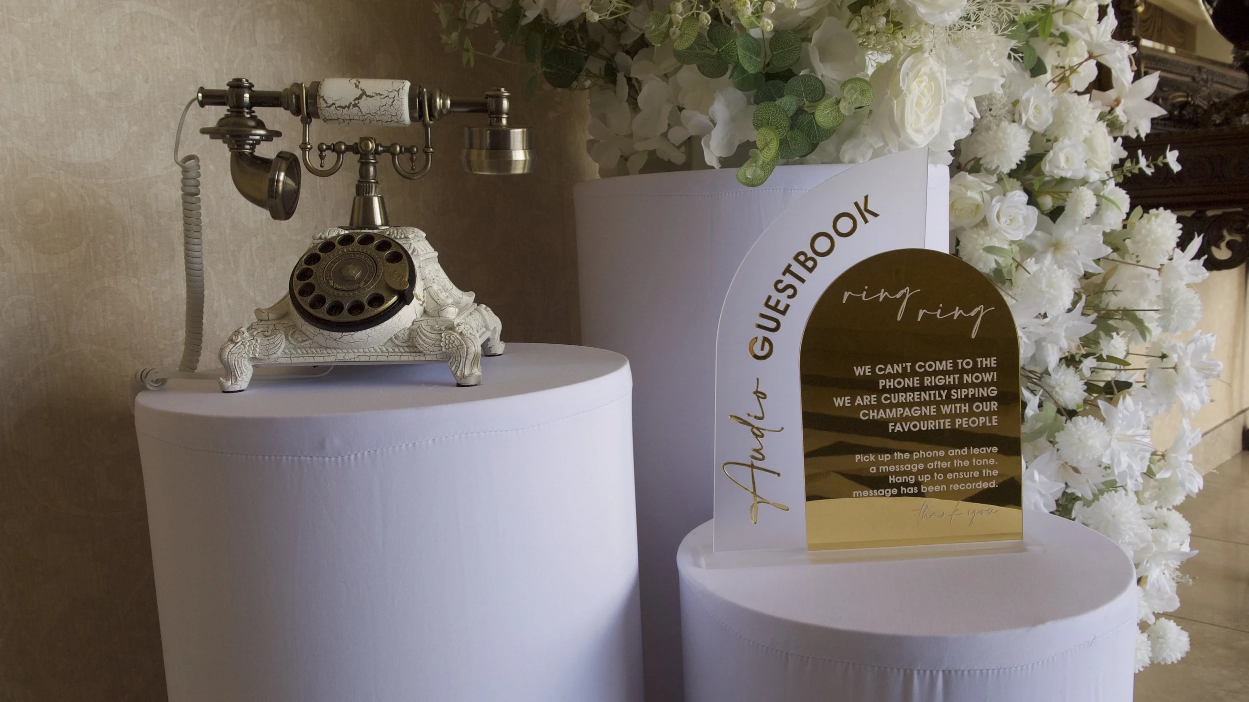 Vintage rotary telephone on a white cylindrical stand near a guestbook sign with white flowers in the background.
