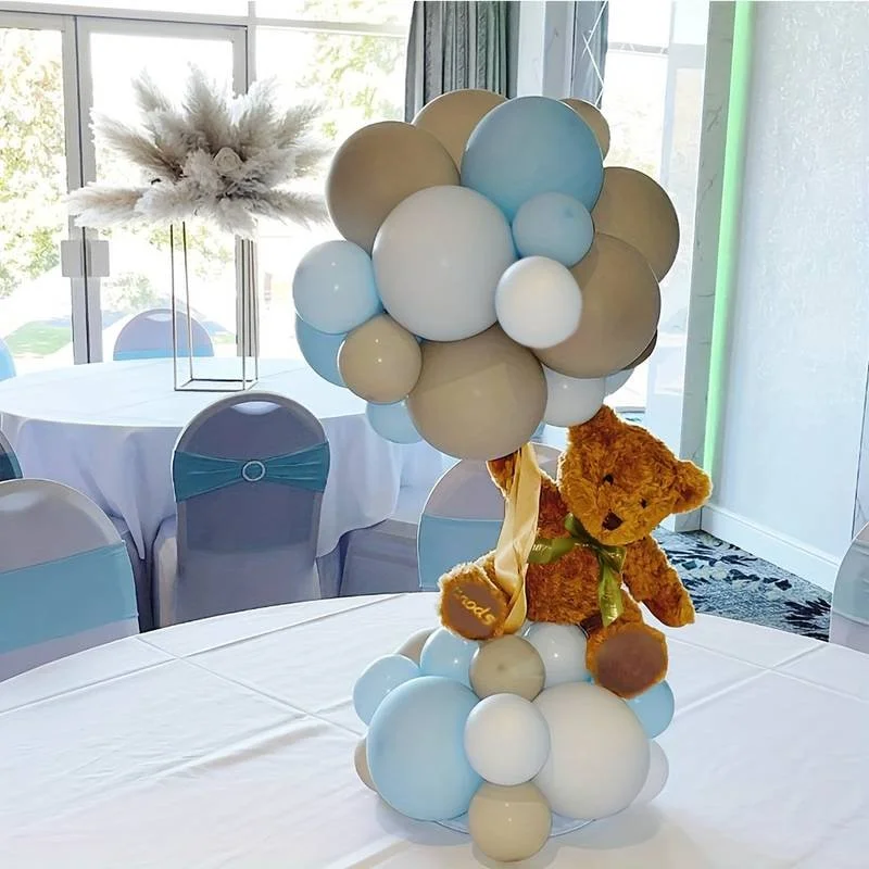A decorative balloon arrangement featuring blue, beige, and white balloons, with a plush teddy bear sitting amidst the balloons. The teddy bear holds a small balloon with the word 'Soon' on it. The arrangement is placed on a table in a decorated event space with round tables and chairs.