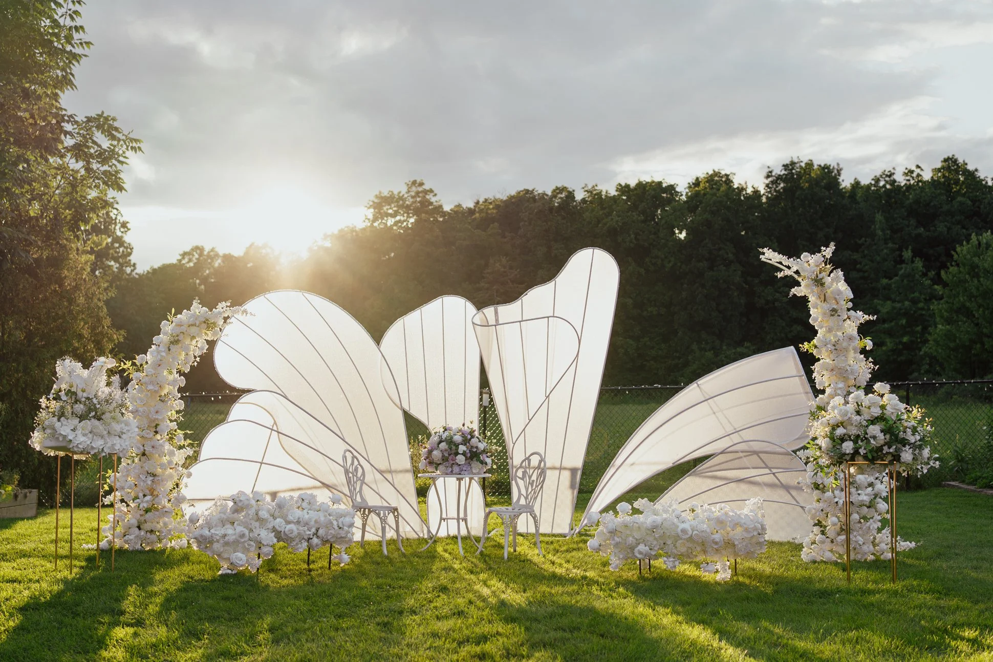 Outdoor wedding ceremony setup with large white backdrop, floral arrangements, and chairs on a grassy lawn at sunset.