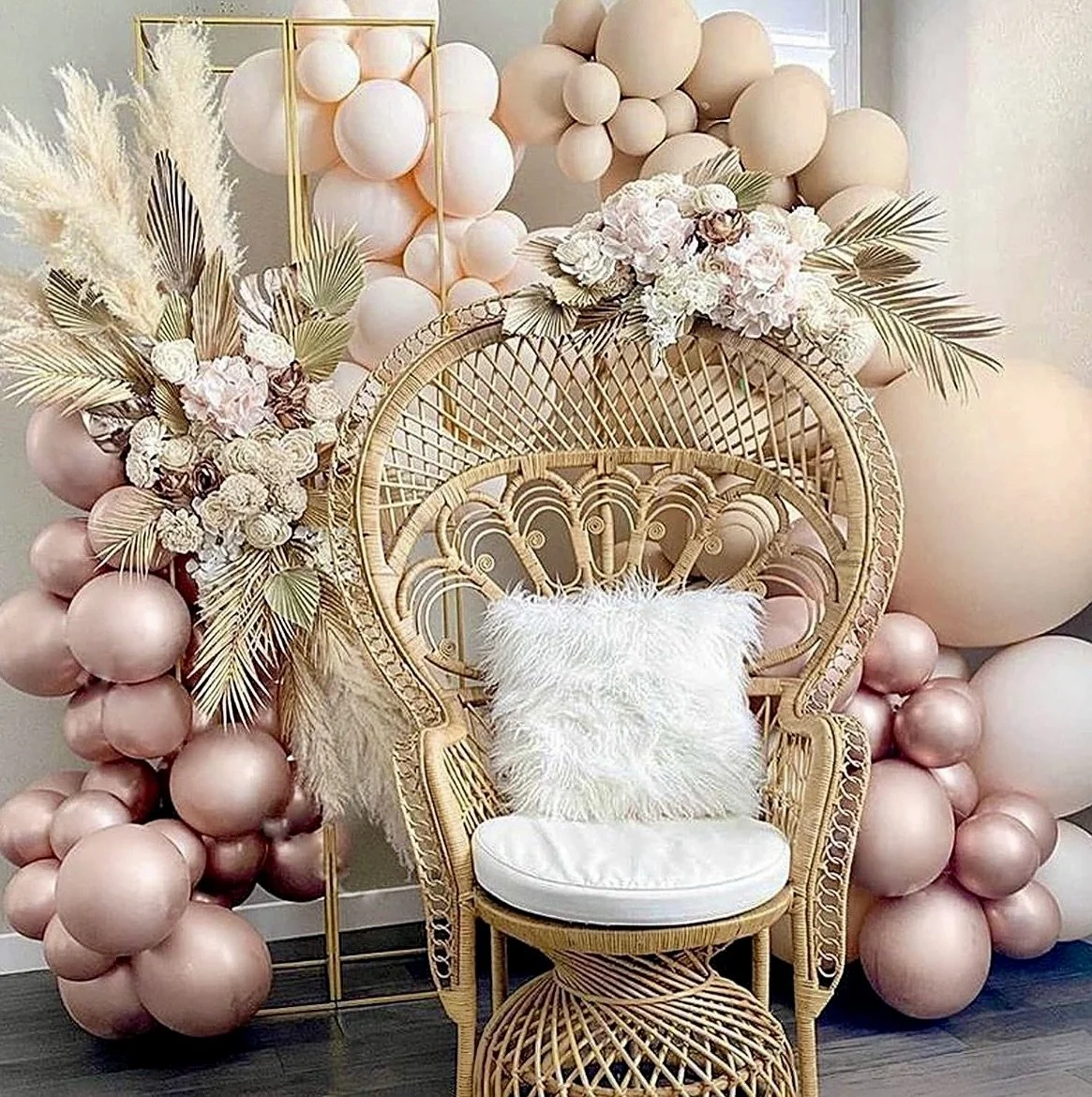Decorative setup with a wicker peacock chair and white cushion, surrounded by beige and pink balloons, pampas grass, and floral arrangements.