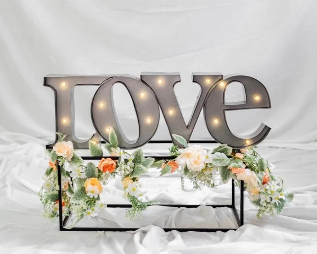 Decorative "Love" sign with lights and floral arrangement on white fabric background.