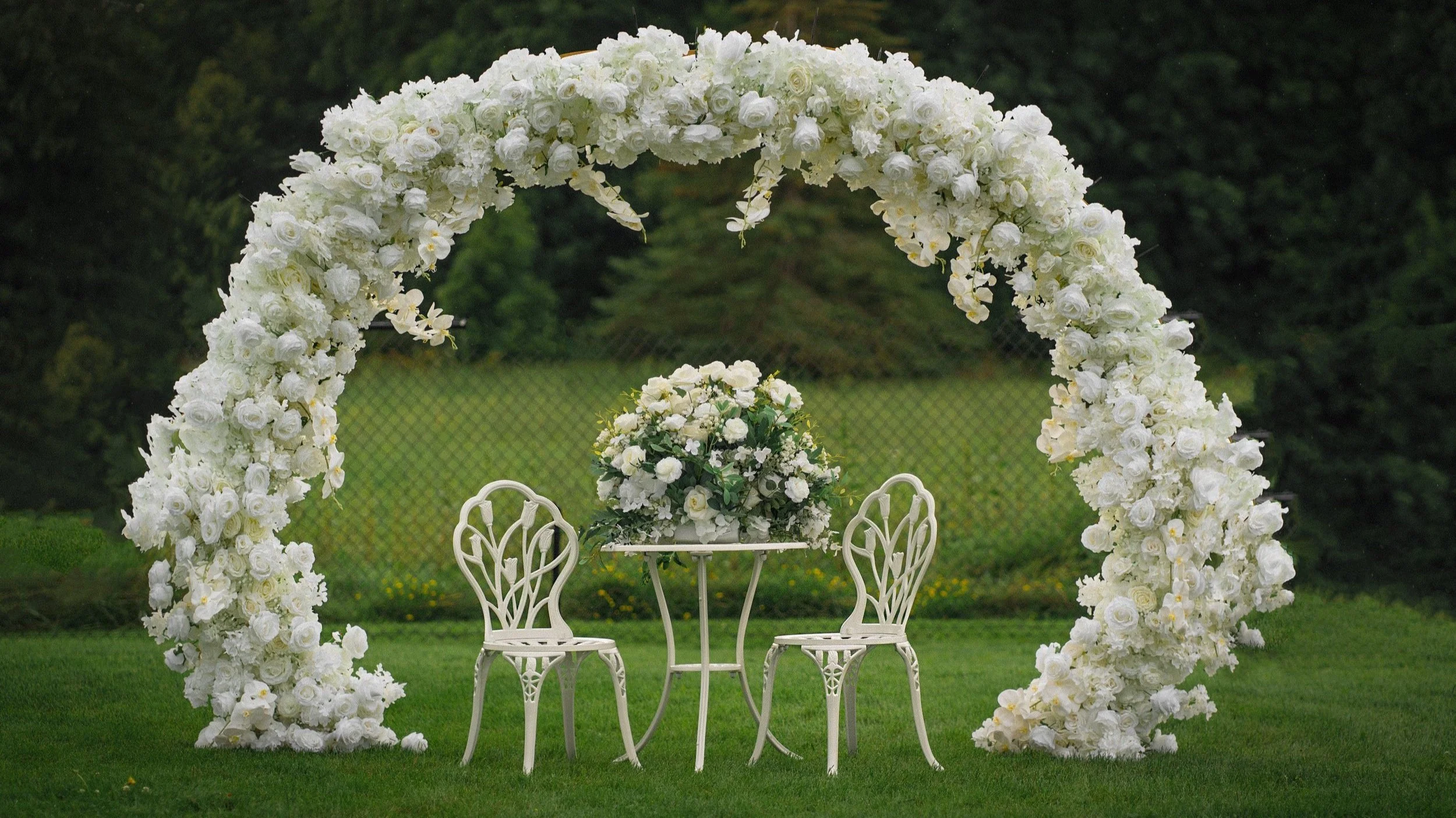 White floral arch with two chairs and a table on grass, decorated with white flowers, against a green outdoor background.