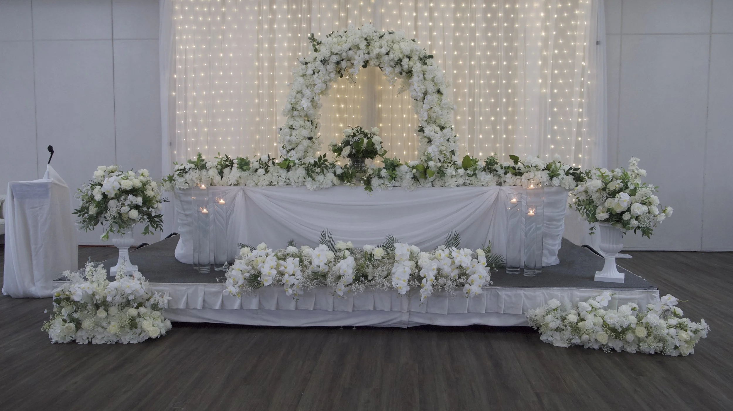 Wedding reception stage decorated with white flowers, drapery, and fairy lights.