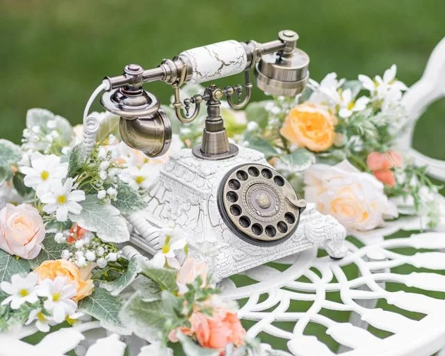 Vintage rotary telephone with white and gold design, surrounded by pink and white flowers on a white decorative surface.