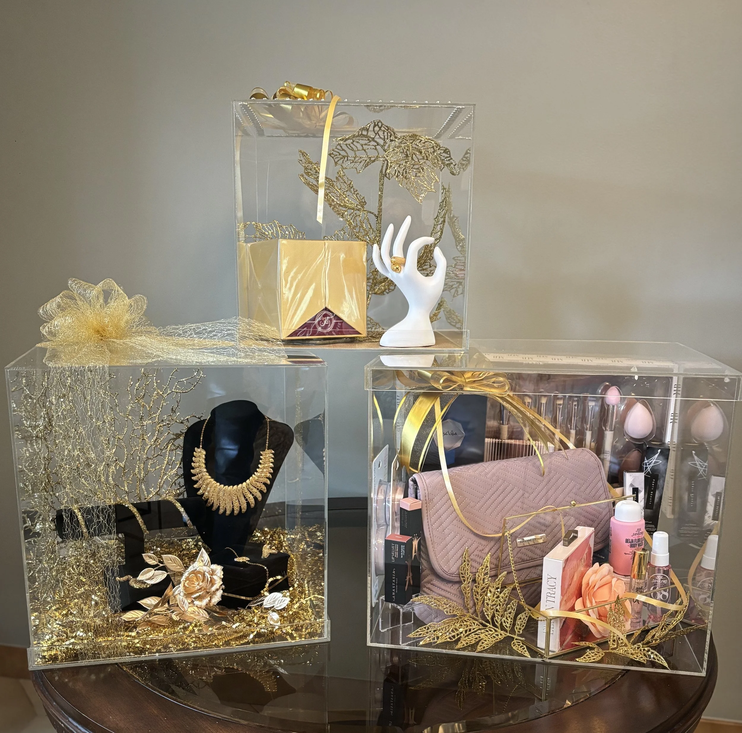 Three transparent display boxes containing decorated gifts. The top box has a gold gift bag, a white hand statue holding a ring, and gold accents. The box on the left features a gold necklace on a black bust, surrounded by gold decorations and flower