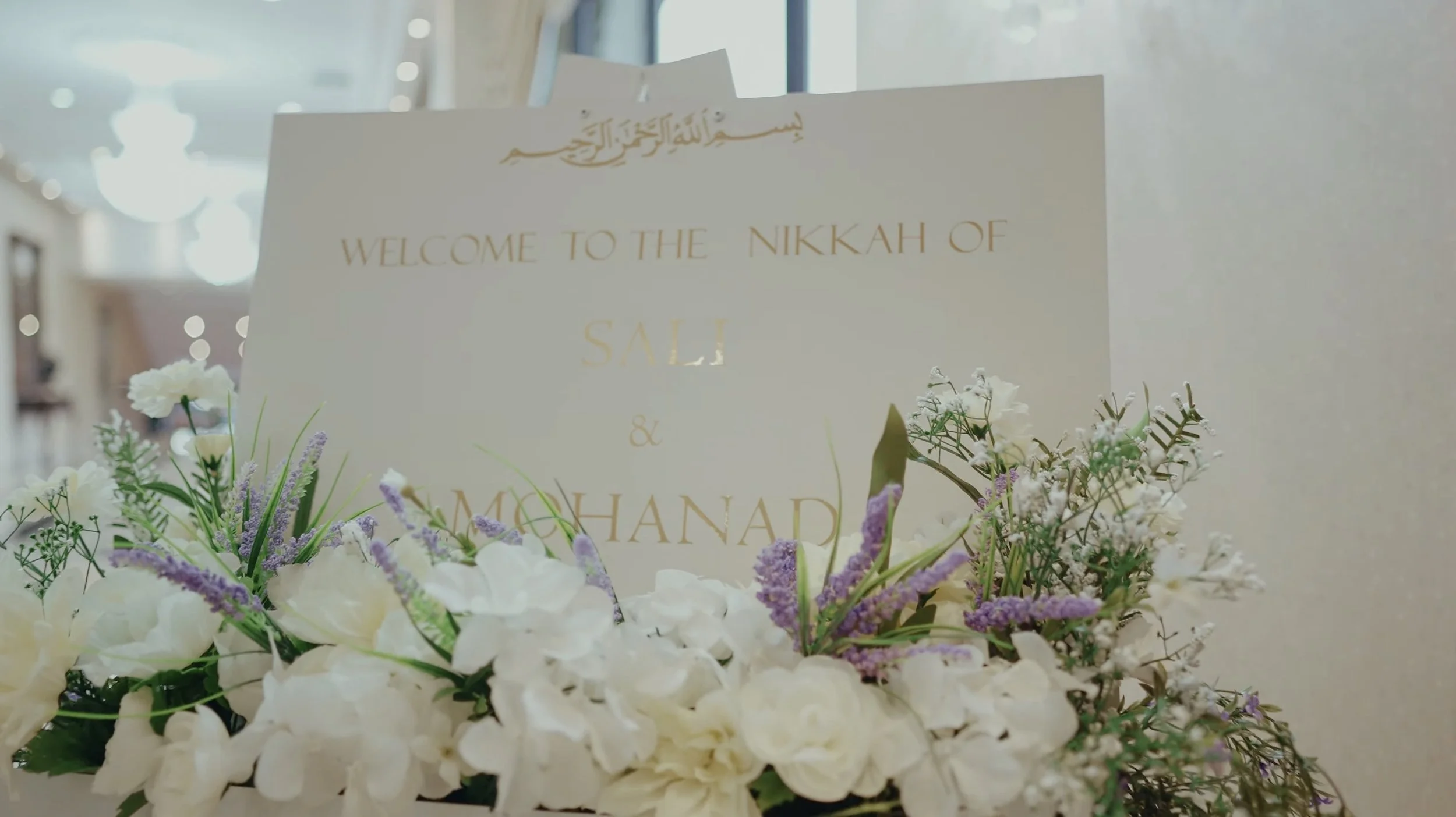 Wedding sign with Arabic greeting, welcoming guests to the nikkah of Sali and Mohanad, surrounded by white and purple flowers.