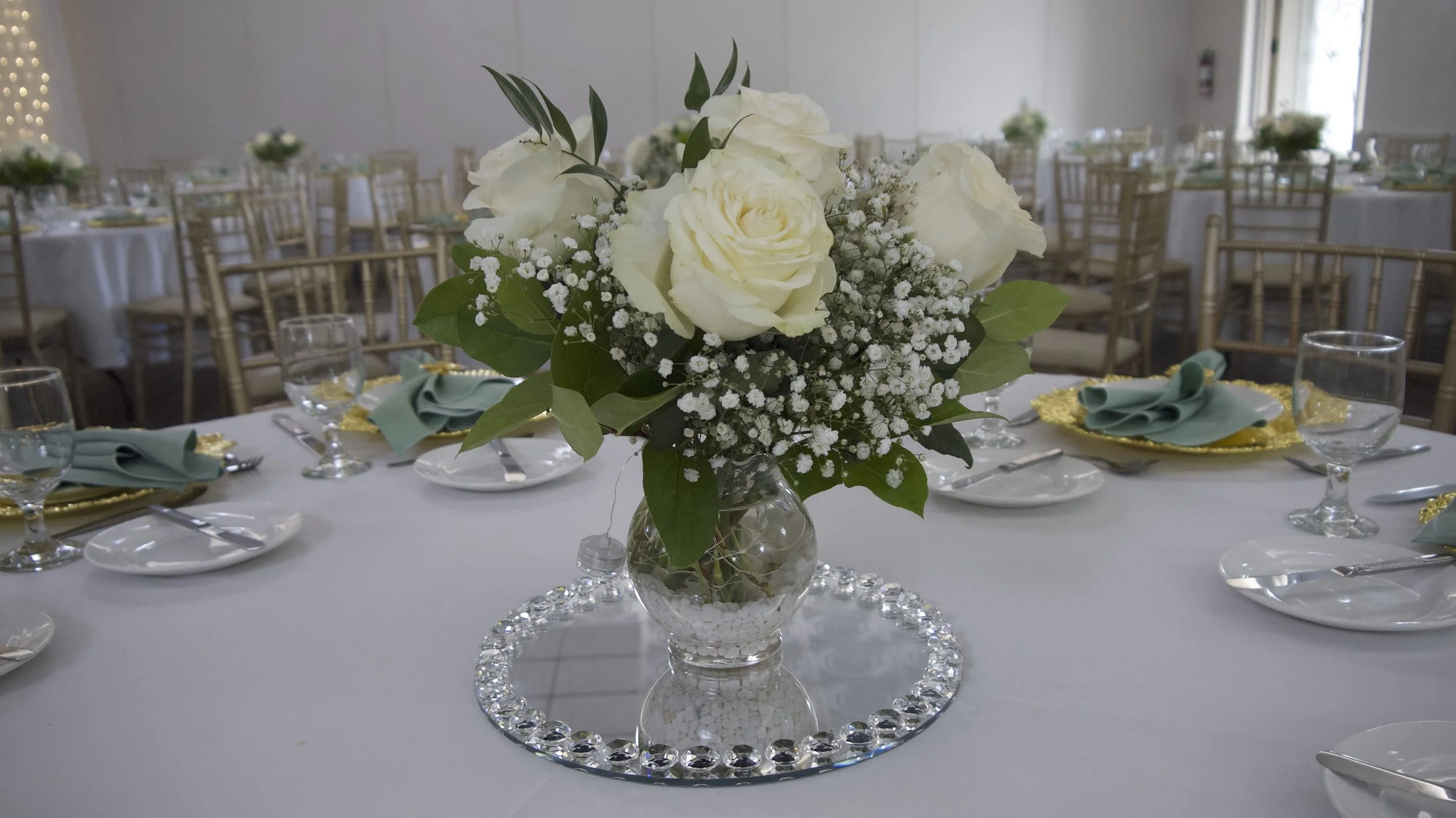 Elegant table setting with a floral centerpiece featuring white roses and greenery, surrounded by glassware and plates on a round, white tablecloth at a formal event venue.