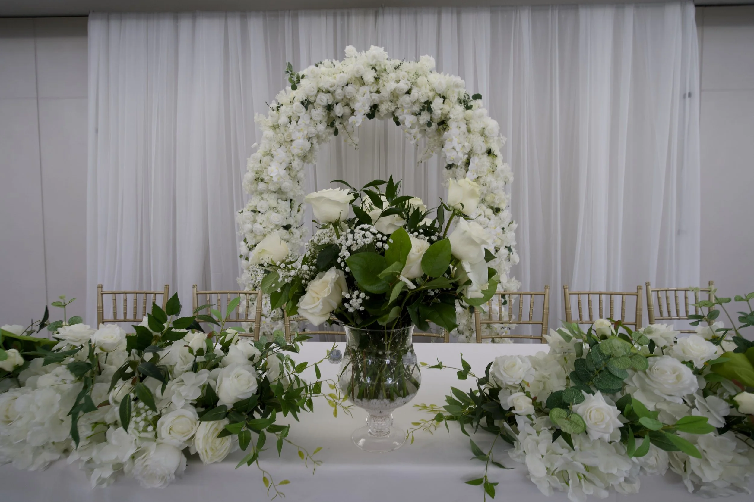 Elegant white floral arrangement with roses and greenery on a table, featuring a large circular flower arch in the background, set against white draped fabric.