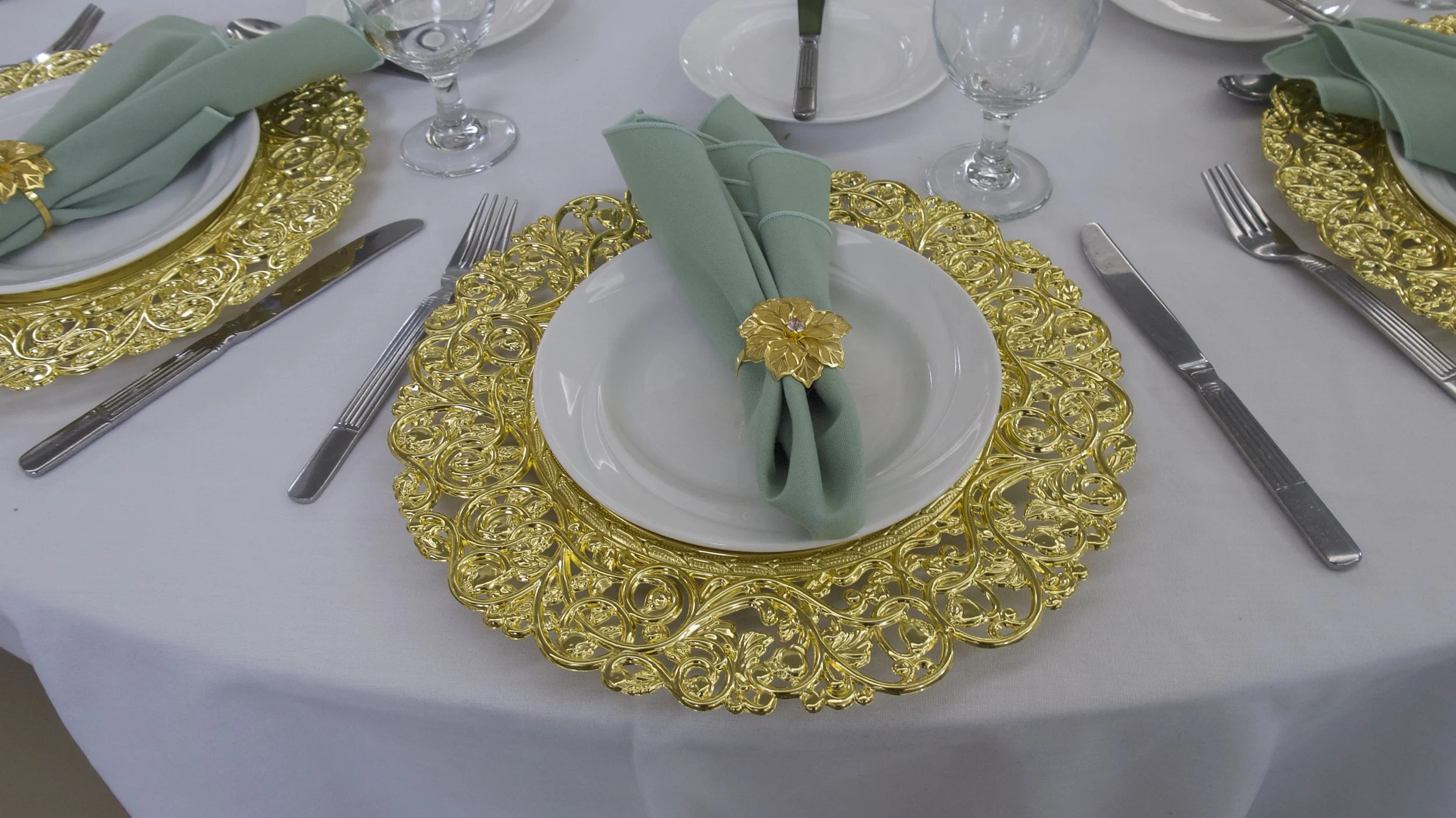 Elegant table setting with gold ornate placemats, white plates, sage green napkins with gold floral napkin rings, and silver utensils on a white tablecloth.