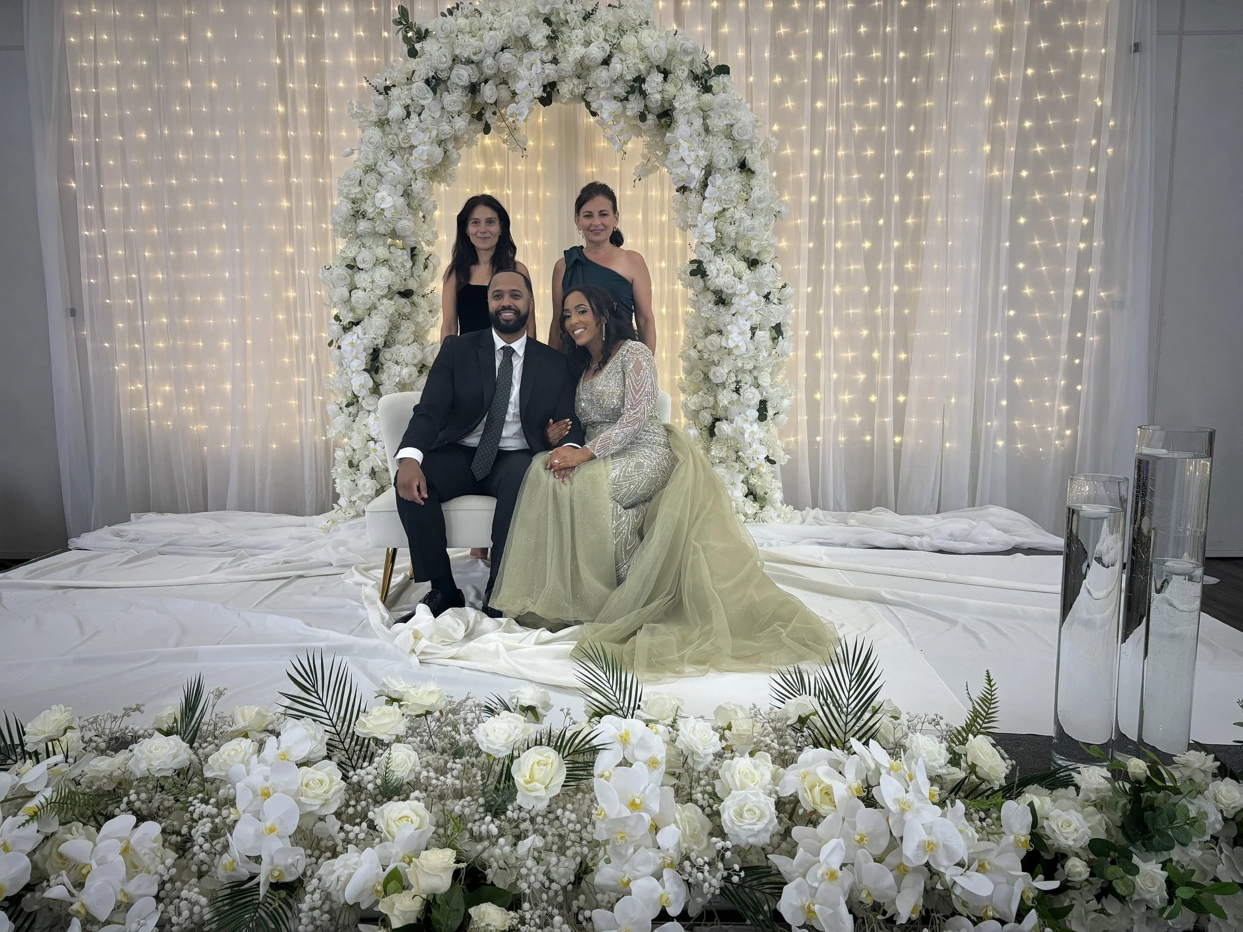 Bride and groom seated on a white chair under a floral arch with white flowers and greenery, surrounded by bridesmaids, with a backdrop of string lights and draped fabric, and floral arrangements in the foreground.