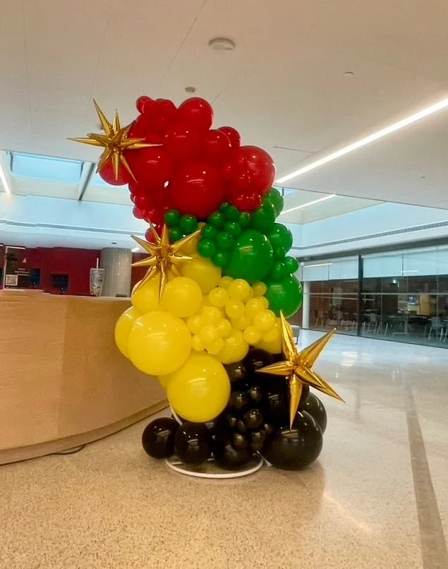 Colorful helium balloon arrangement in red, green, yellow, and black with gold star-shaped balloons, set up indoors near a reception desk.