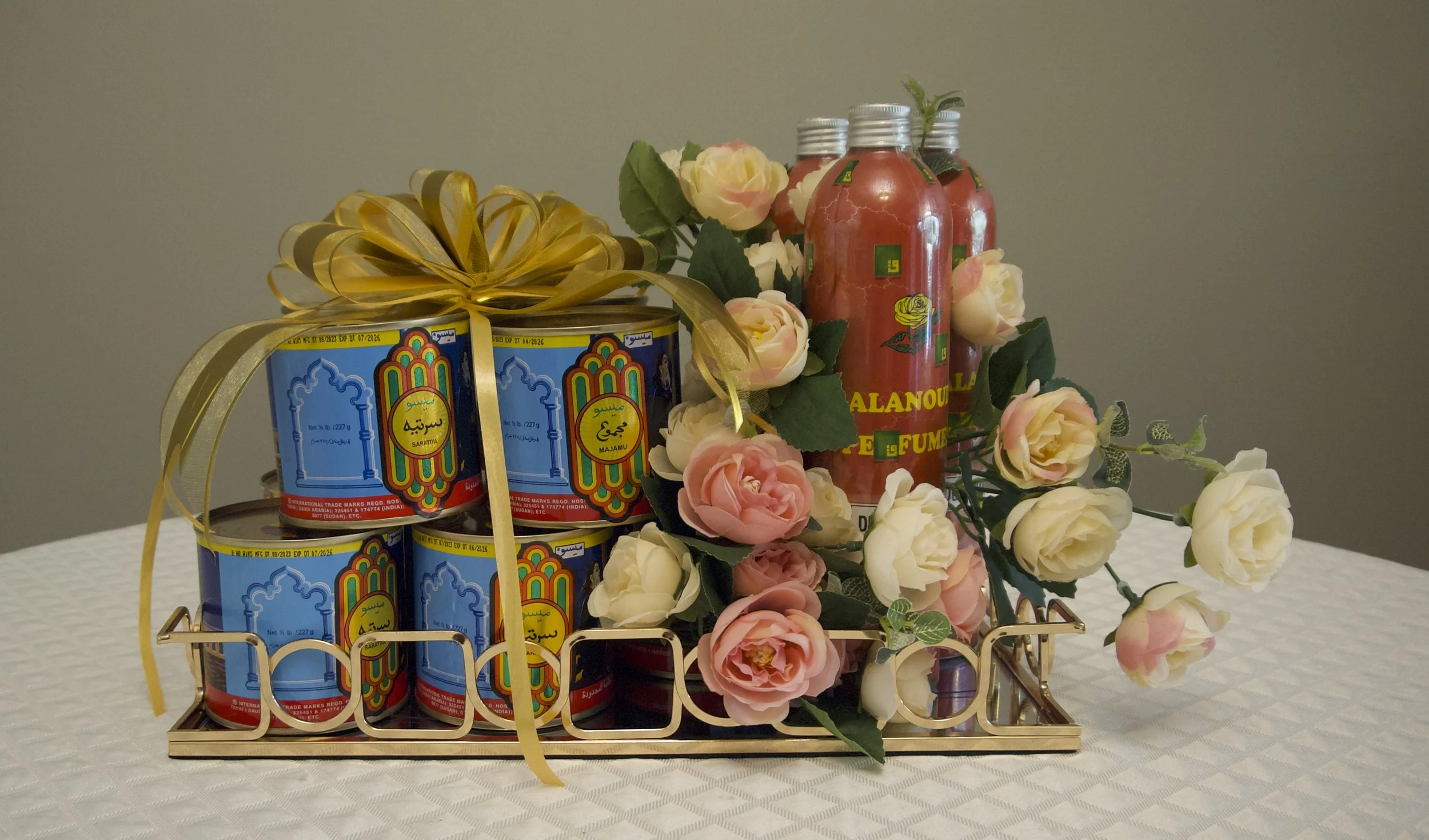 Gift basket with cans of Saratya preserved food, four bottles of flavored drink, and artificial pink and white roses on a white tablecloth.