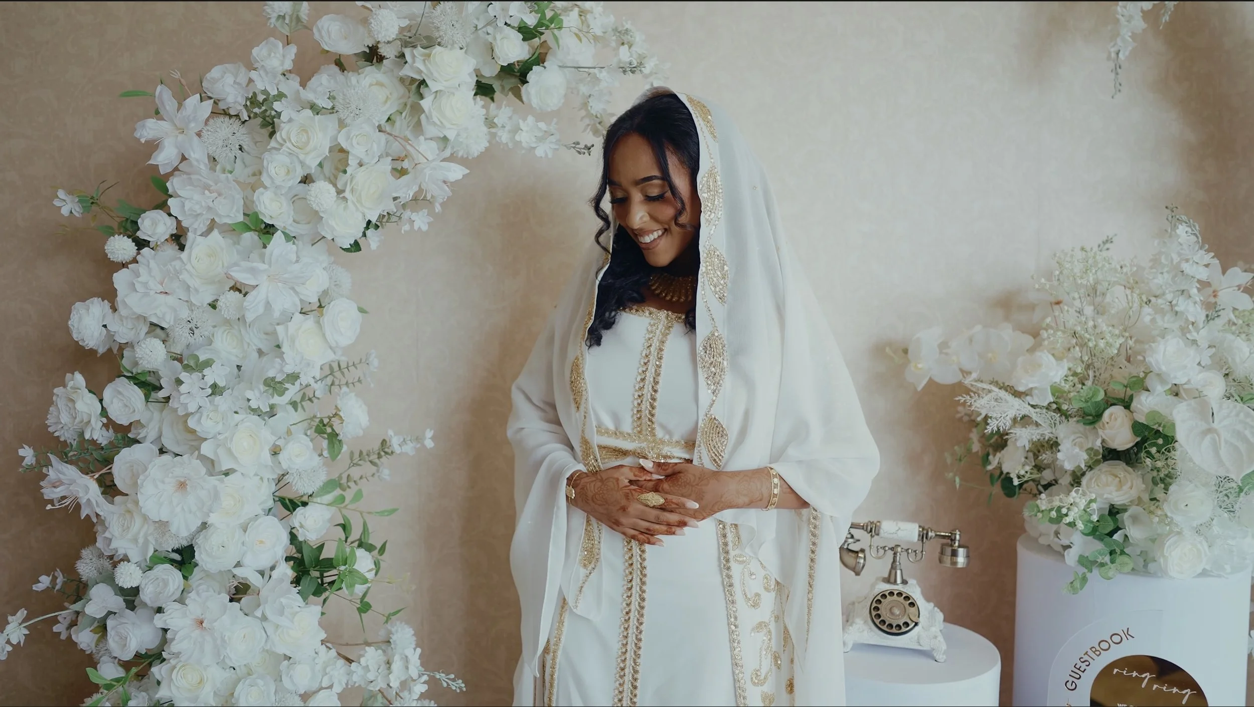 Bride dressed in white traditional attire standing amidst white floral decorations and a vintage telephone.