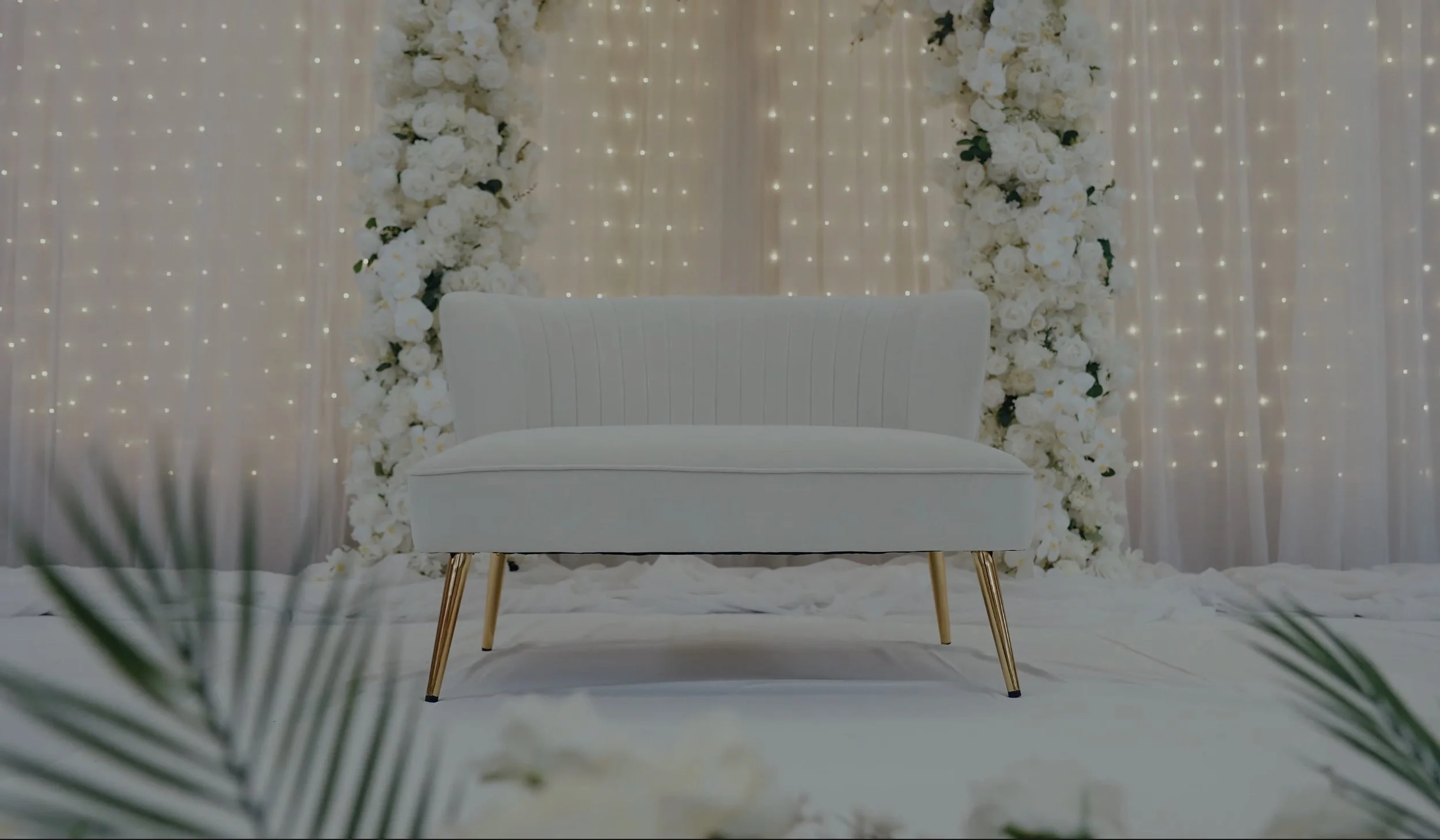 White couch with gold legs in a decorated setting with white floral arrangements and string lights.