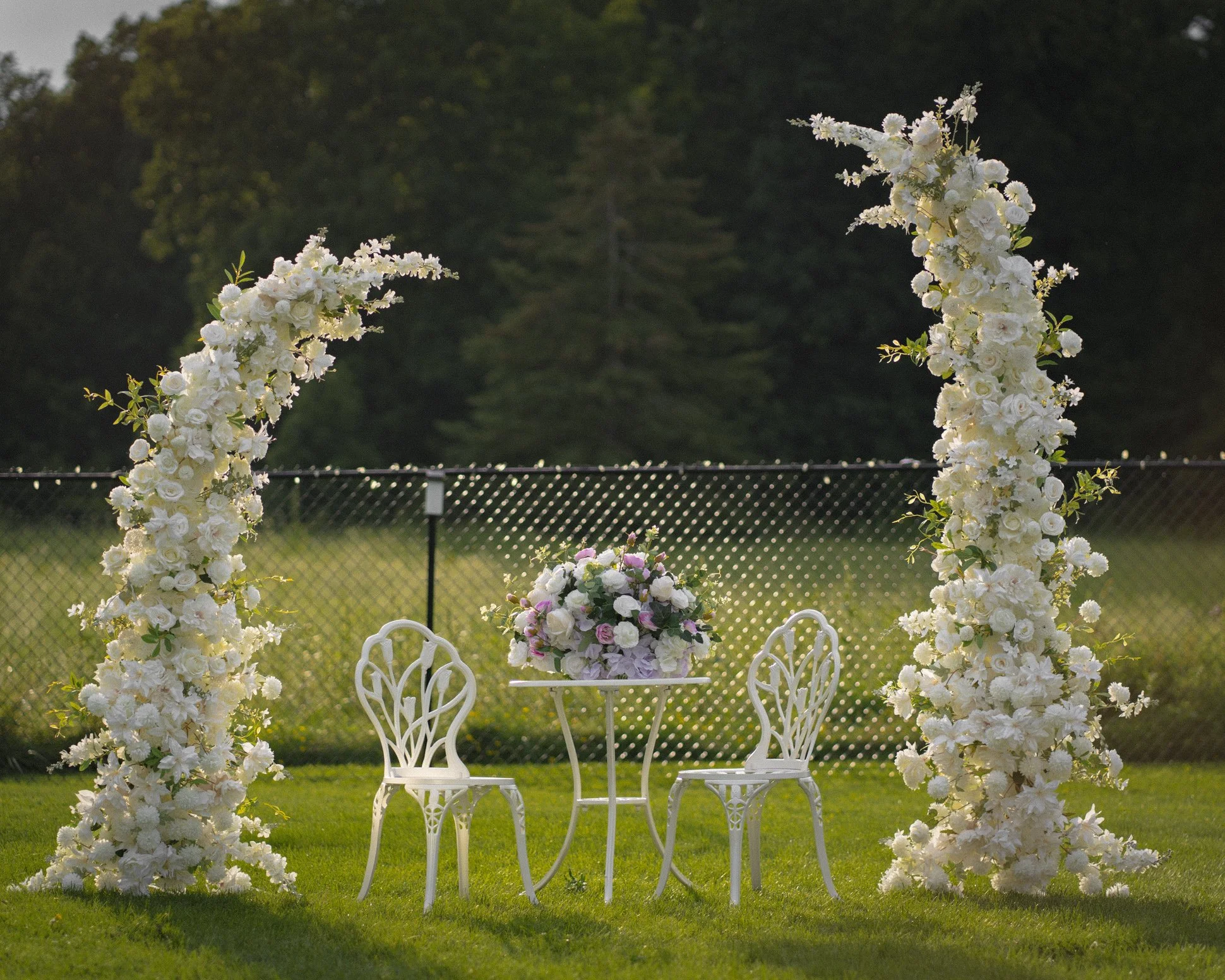 Outdoor setting with two white chairs and a table adorned with a floral arrangement, flanked by two large floral arches made of white flowers, on a grassy area with trees and a fence in the background.