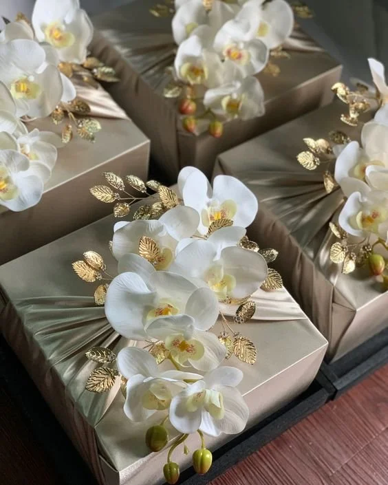 Elegant gift boxes with beige wrapping, adorned with white orchids and gold leaves, arranged neatly.