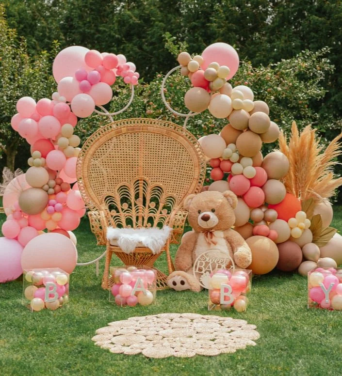 Decorative setup for a baby shower or celebration with pink, beige, and peach balloons, a wicker chair with fluffy white cushion, a large teddy bear holding a