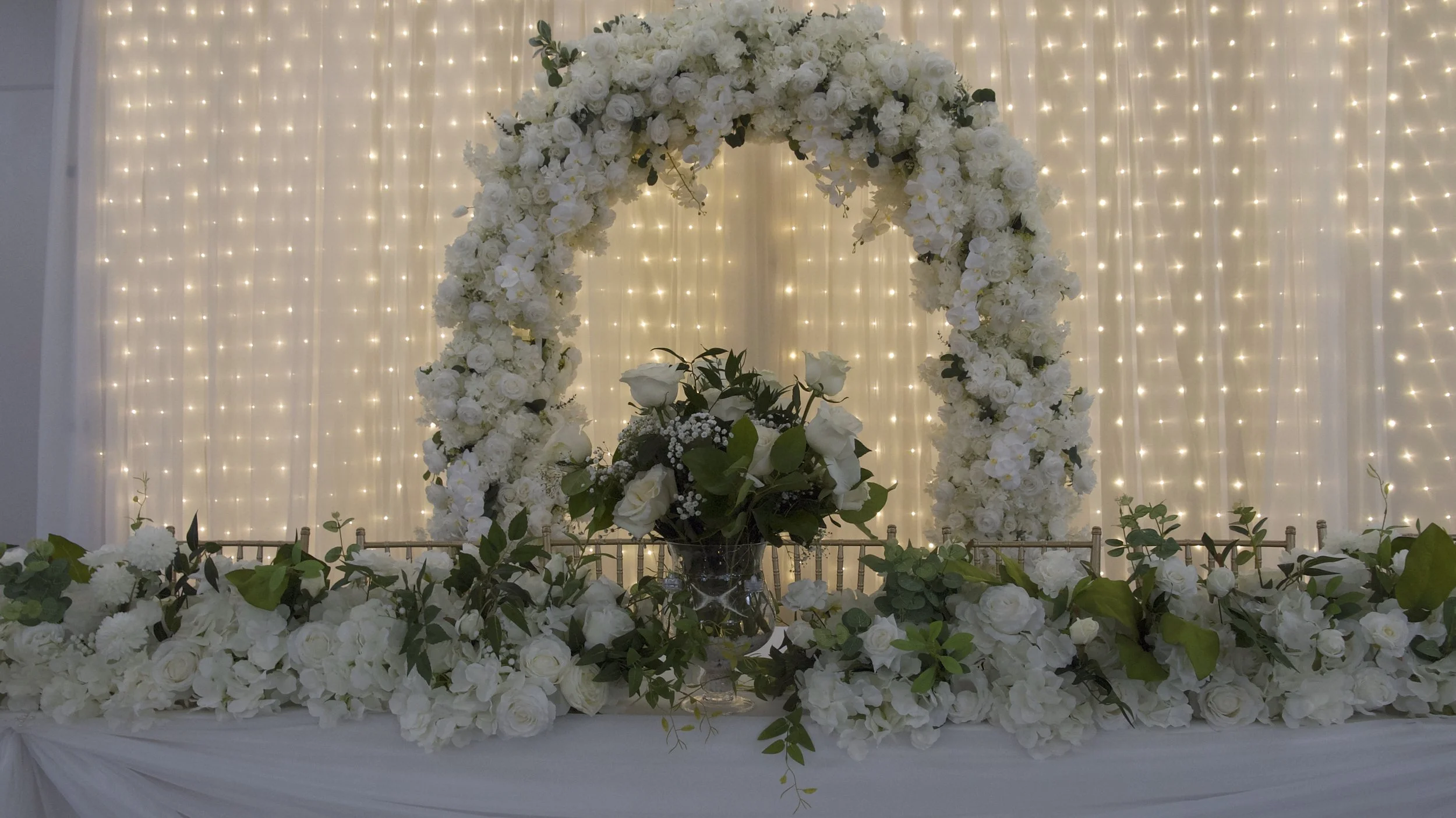 Floral wedding arch with white roses and greenery, illuminated by string lights.