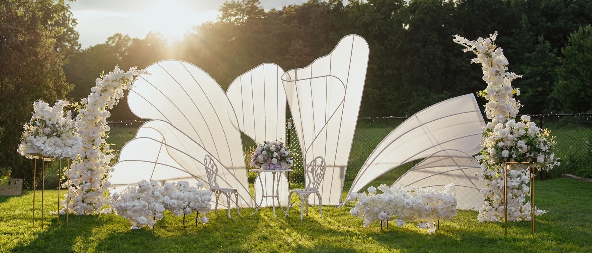 Outdoor wedding ceremony setup with white floral arrangements, chairs, and decorative panels on a grassy area.