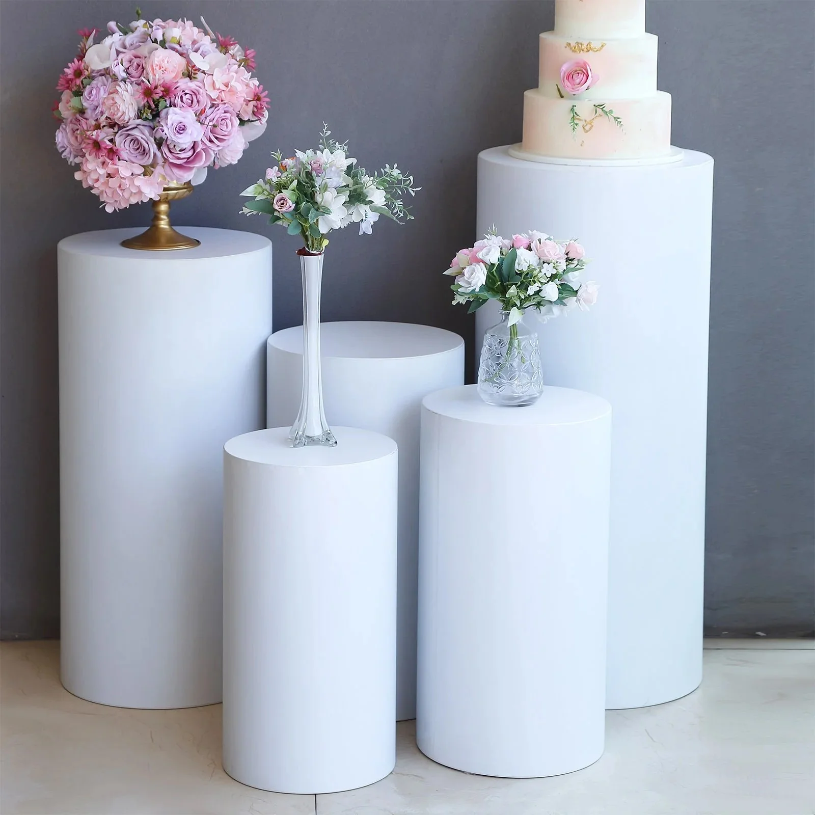 White cylindrical pedestals with floral arrangements and a four-tier wedding cake against a gray background.