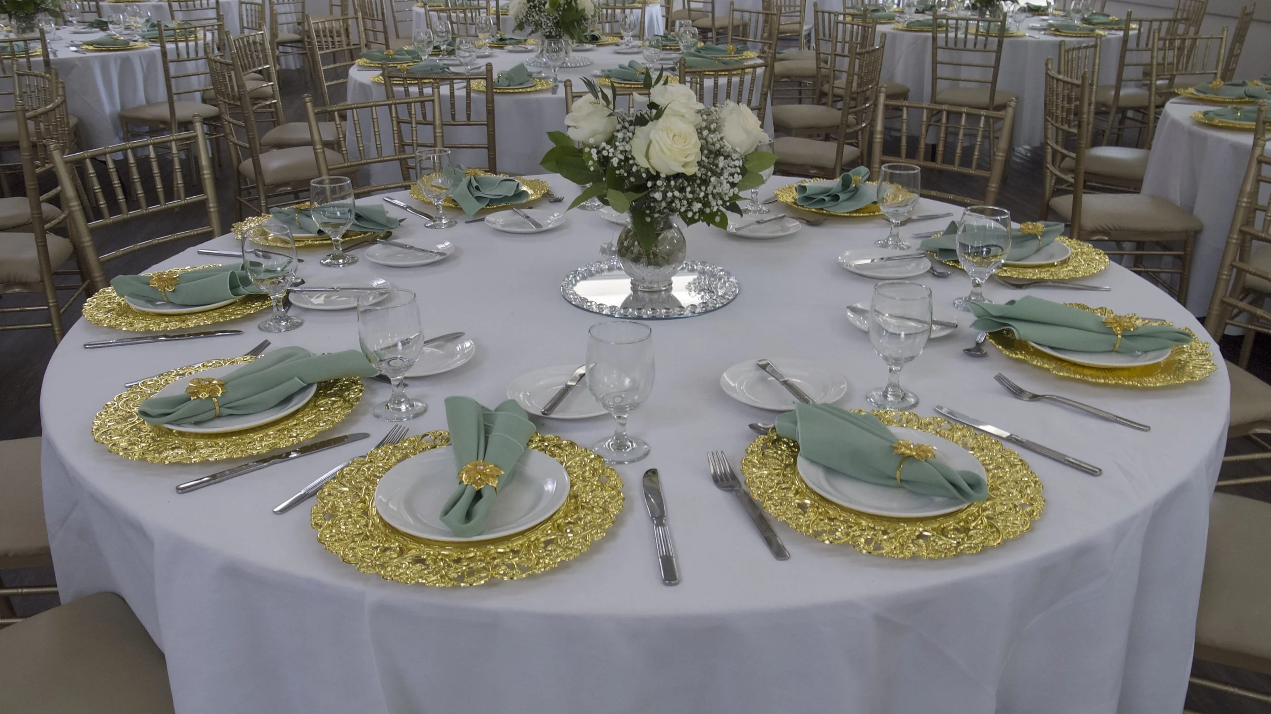 Elegantly set round banquet table with white tablecloth, gold placemats, green napkins with gold rings, white dishes, and glassware. Centerpiece of white roses and greenery. Surrounded by gold chairs.